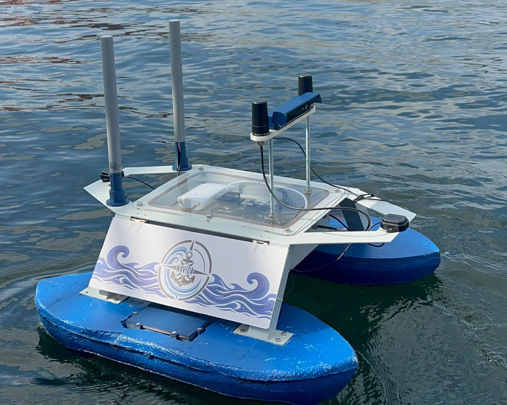Team Navi’s unmanned surface vessel afloat in Trondheim harbour — twin blue catamaran hulls, a white instrument deck carrying the stereo camera housing, and two GNSS antenna masts.